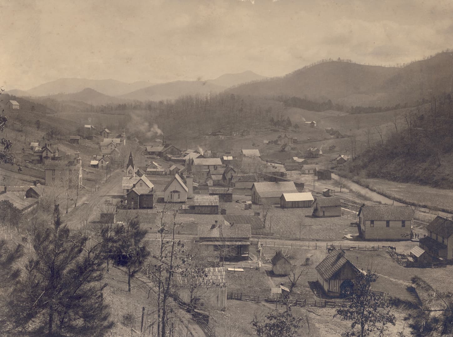 Sepia-toned historic wide-angle photograph of a small mountain town in a valley, with a prominent white church and steeple, a dirt road, and surrounding farmland and forested hills.