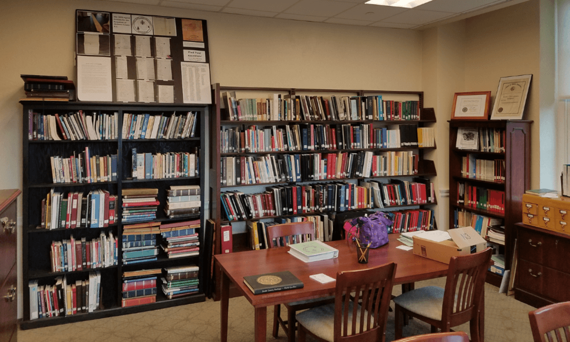 Color photograph of the interior of the JCGS research library, showing bookshelves full of genealogy and reference volumes, a reading table with chairs, framed certificates on the wall, and a card catalog cabinet.
